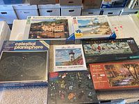 Seven sealed jigsaw puzzles arranged on a surface. Includes Ravensburger puzzles with colorful scenic art, Celestial Planisphere puzzle and others with natural and thematic images.