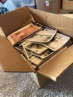 Open cardboard box filled with a large stack of old photos, showing top edges of many black and white and sepia photographs.