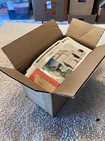 Open cardboard box filled with stacks of old photographs with one visible on top showing a person next to a machine or vehicle.