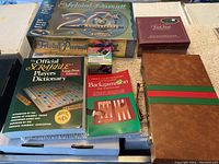 Top view showing Trivial Pursuit 20th Anniversary Edition box, Official Scrabble Players Dictionary, Backgammon game, brown book-style case, and a sealed boxed item.