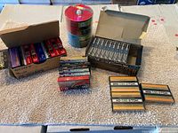 Photo showing multiple boxes and stacks of unopened and unused blank audio cassettes and stacks of CDs/DVDs on spindles.