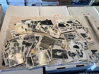 Large pile of vintage black and white photographs spread on a beige textured cloth on cardboard boxes. Photos show various people and scenes.