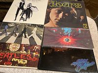Top-side front covers of six classic rock vinyl records featuring Fleetwood Mac's Rumours, The Doors self-titled, Abbey Road by The Beatles, Sgt Peppers Lonely Hearts Club Band by The Beatles, Santana's Abraxas, and Hotel California by The Eagles.