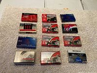 Top-down photo of all 12 sealed cassette tapes laid out on a textured surface showing variety of brands and tape lengths.