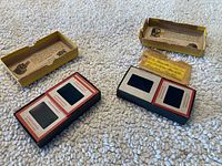 Two photographic slides in black and red mounts with clear viewing windows resting on a white textured surface along with two small open cardboard slide storage boxes, one with a handwritten mailing label.