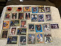 All cards laid out on a textured white surface, displaying the full assortment of 27 cards in plastic cases, showing basketball, baseball, and hockey players.