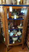 Front view of antique wood display cabinet with glass door open, showing blue shelves with various decorative items inside.