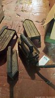 Three antique Koenig gas irons and three matching metal resting bases arranged on a wooden floor covered with worn plywood sheets. The irons have a dark cast iron body with metal handles and show rust and dirt.