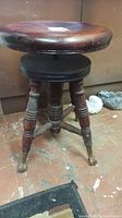 Full view of antique wooden piano stool with adjustable round seat, turned legs and claw feet with glass balls. Shows surface scuffs and wear.