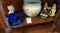 Wide view of lot showing two ceramic cat piggy banks, two cobalt blue glass candle holders, floral planter, house and tree pitcher, and porcelain figurines.