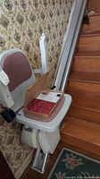 Full view of stairlift chair fixed to stair rail with pink cushioned seat and backrest, situated on wooden staircase with floral wallpaper and carpet