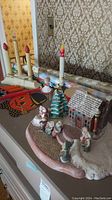 View of vintage Christmas village with painted ceramic house featuring colored lights, figurines including unattached Santa, electric candles, Halloween-themed wrapping paper, and ribbons on surface.