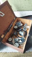 Open wooden box showing assorted vintage gauges of different types and sizes inside, along with cables and connectors.