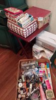 Full view of sewing baskets on a red folding table with packing boxes and open box of sewing supplies on the floor.