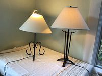 Two metal table lamps side by side on a table, both with black metal bases and cream shades, highlighting different base shapes.