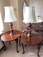 Two red stained oval wooden side tables with folding sides, each with a brass table lamp placed on top.