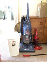 Full shot of the three items: upright Bissell vacuum in blue, red Dirt Devil hand vacuum, and white Rubbermaid waste basket.