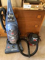 Photo showing both the Bissell upright vacuum and the Shop-Vac side by side on carpeted floor with wooden furniture in the background.