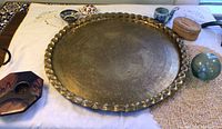 Large round brass platter with scalloped edge and etched pattern laying flat on table with other items visible