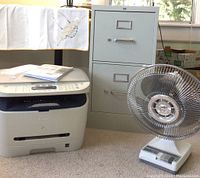Photo showing Canon multifunction printer, 2-drawer metal file cabinet, and desktop fan on carpeted floor.