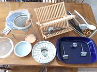 Overview of multiple kitchen tools and bakeware on a wooden table, showing Fiesta Ware bowl, baking pans, thermometers, wooden dish rack, and utensils.