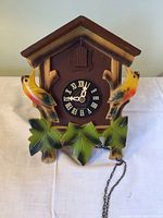 Front view of wooden cuckoo clock featuring painted birds and leaves, black Roman numeral clock face, and visible metal chain.