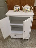 Front view of white medicine cabinet open, showing shelves and drawer, with floral ceramic teapot and planter on top