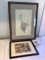 Framed monochrome botanical print leaning against the wall, smaller Chinatown print laid flat on table along with small ceramic piece.