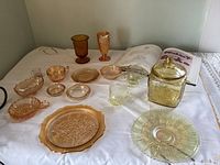 Collection of iridescent glassware items including a large yellow lidded jar, goblets, cups, plates, and dishes in various shapes and sizes spread on a white cloth.