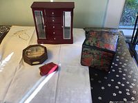 Photo shows a tabletop with three jewelry boxes: a reddish brown wooden chest with glass-paneled side doors and multiple drawers, an octagonal jewelry box with a clear top and sectioned compartments, a black lacquered jewelry box with colorful patterns. A small red change purse and black wall hanging are also visible.