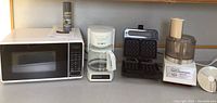 Overview photo of the four kitchen appliances on countertop: microwave oven, drip coffee maker, waffle maker, and food processor with blade removed.