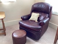 Brown leather Barca Lounger recliner chair with matching round footstool placed in a carpeted corner near windows and a small wooden table. Shows visible leather wear and creases.