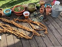 Photo showing tan macrame plant hangers laid out on a wood deck alongside various ceramic and plastic pots, some containing moss and green plants.