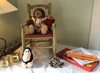 Full shot of doll rocking chair with two stuffed bears seated on it, plush penguin and striped plush on table, games, and votive candle holders.