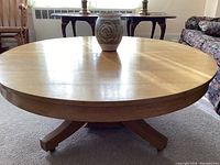 Front view of the round wooden coffee table showing the tabletop and pedestal base with wheels, with pottery vase centered on the table.