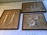 Three framed fabric art pieces inspired by sandstone petroglyphs, showing human and animal figures on brown textured fabric backgrounds.