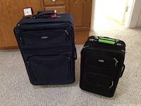 Photo showing two black soft suitcases of different sizes placed side by side and a small travel alarm clock on top of the large suitcase.
