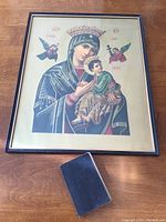 Framed religious print showing Madonna and Child with crowns and angels, accompanied by a vintage New Testament book with dark cover on wooden floor