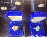Top-down image of various rings and pins displayed in dark velvet holders and blue velvet boxes, showing silver ring with blue glass center, silver ring with turquoise stones and several pins.