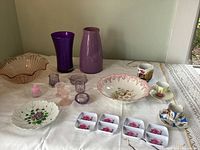 Photo showing entire assortment of vintage glass and porcelain items laid out on a white tablecloth. Items include purple glassware, pink scalloped bowl, porcelain cups and saucers, white plastic dipping trays with pink roses, and decorative plates with floral motifs.