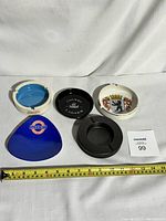 All five assorted advertisement ashtrays arranged with measuring tape for scale, showing variety in shape and color.