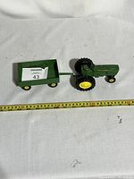Side view of green John Deere diecast metal tractor connected to pull trailer, placed on white surface with measuring tape for scale showing approximately 12 inches in length.