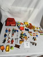 Overview photo showing assortment of plastic toys and figures laid out including multiple Fisher-Price and Playmobil figures, small toy animals, furniture pieces, wagons and carts, and a red carrying case.