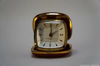 Front view of the open clock showing the square white clock face with black numbers 12, 3, 6, 9 and gold tone edges, clock hands, and alarm indicator.