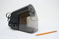 Black and grey X-ACTO SchoolPro electric pencil sharpener with attached power cord and transparent shavings bin, shown next to a standard pencil for scale.
