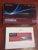 Two Blu-ray players in boxes: Toshiba black box with blue light effects and white Sony box with product label.