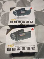 Two boxed HD digital video cameras in sealed factory packaging with printed retail box showing key features and specifications.