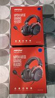 Top side view of two MPOW AirSE Gaming Headsets in sealed factory packaging on a grey patterned surface.