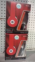 Two SD brand stereo headphones in red and black colors, in factory packaging. Displayed against pegboard background.