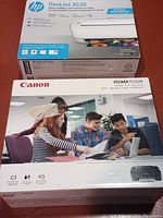 Two boxed printers side by side, HP DeskJet 3630 on top and Canon PIXMA TS3329 below, both in factory packaging.